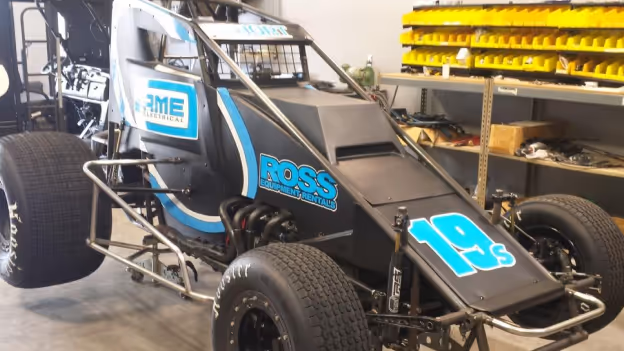 Black sprint car with blue and white decals, including 'ROSS Equipment Rental' and the number 19s, parked indoors near shelves with yellow bins.