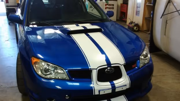 Front view of a blue Subaru car with white racing stripes on the hood in a garage.
