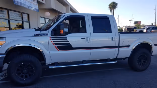 Side view of a white pickup truck with black and red stripe decals parked outdoors in a sunlit area.