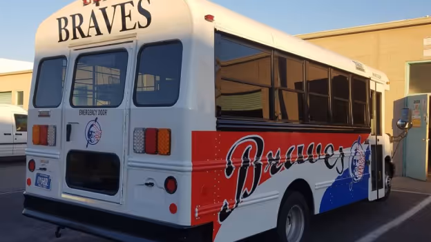 White and red school bus with 'Braves' logo and mascot illustration parked in a lot near a beige building.