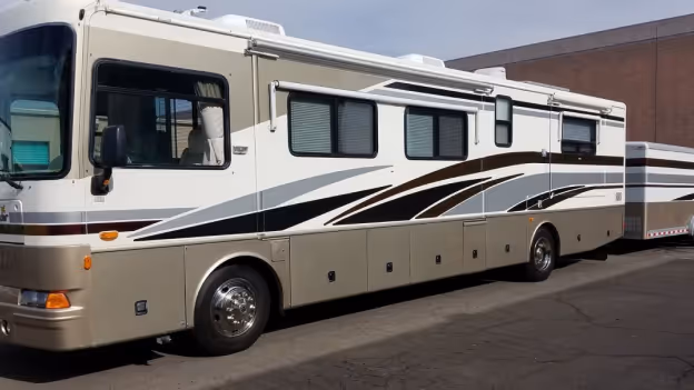 Large beige and white motorhome with black and brown stripes parked on pavement against a building.