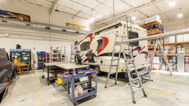 Two technicians working on a white RV inside a large, well-lit garage with ladders and tools around.
