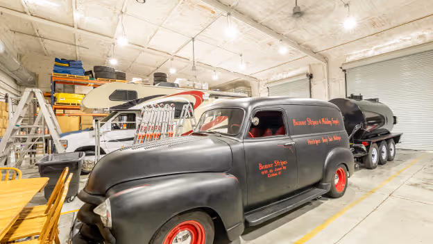 Vintage black delivery truck with red wheels and lettering parked in a spacious warehouse with other vehicles and equipment.