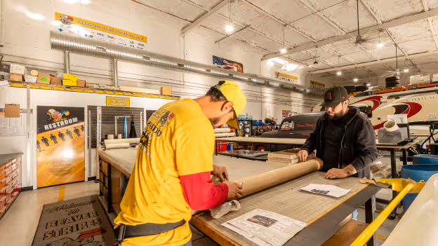 Two men working together rolling out material on a large table inside a busy workshop with tools and equipment.
