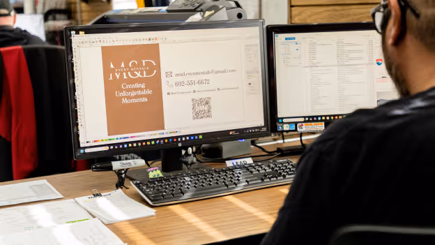 A person working at a desk with two computer monitors, one displaying an event rental advertisement and the other showing an email inbox.