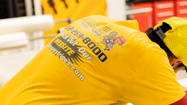 Person wearing a bright yellow shirt and cap with a company logo and website on the back against a workshop background.