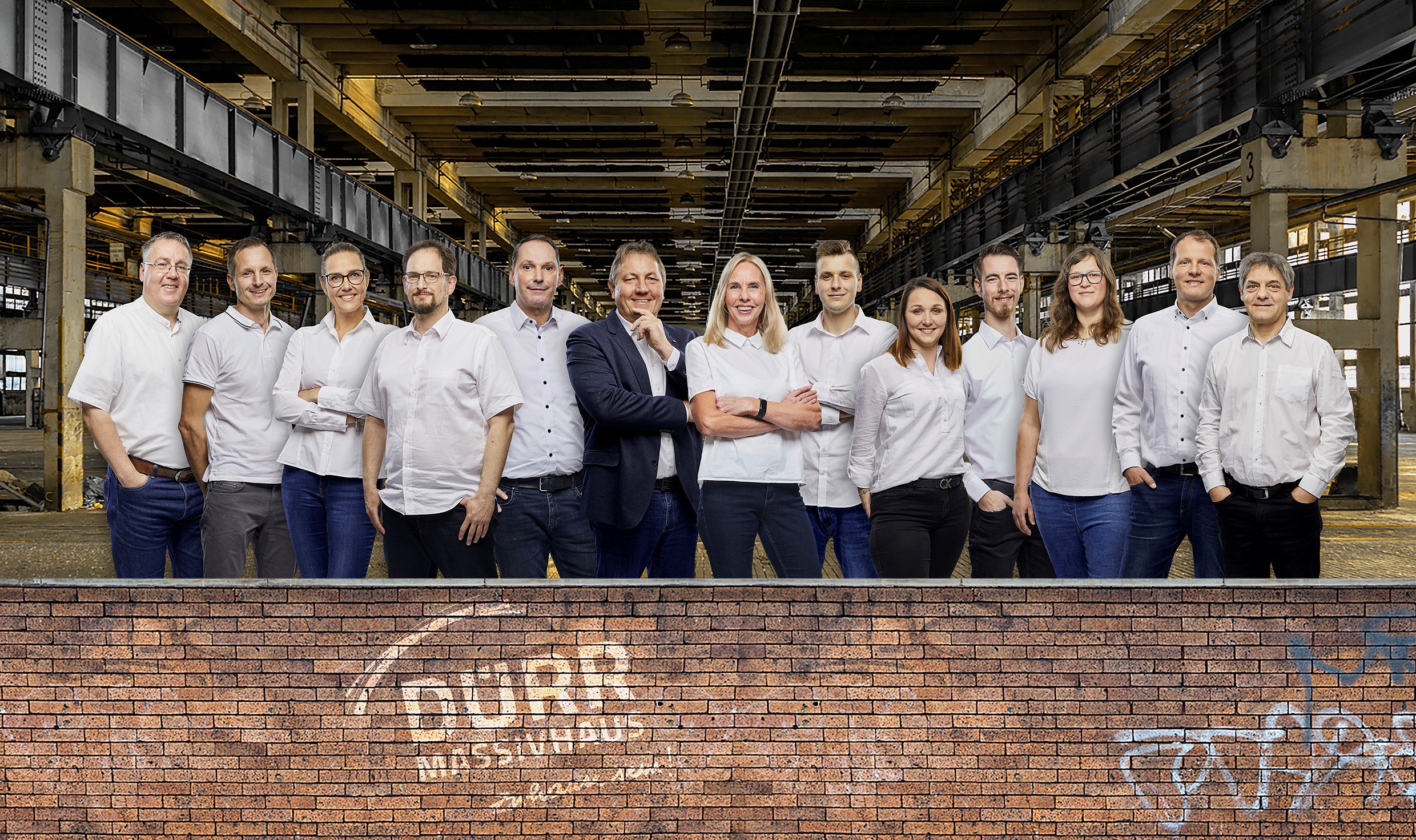Gruppenfoto von zwölf Personen in weißem Hemd und Bluse, die vor einer großen, leeren Industriehalle stehen.