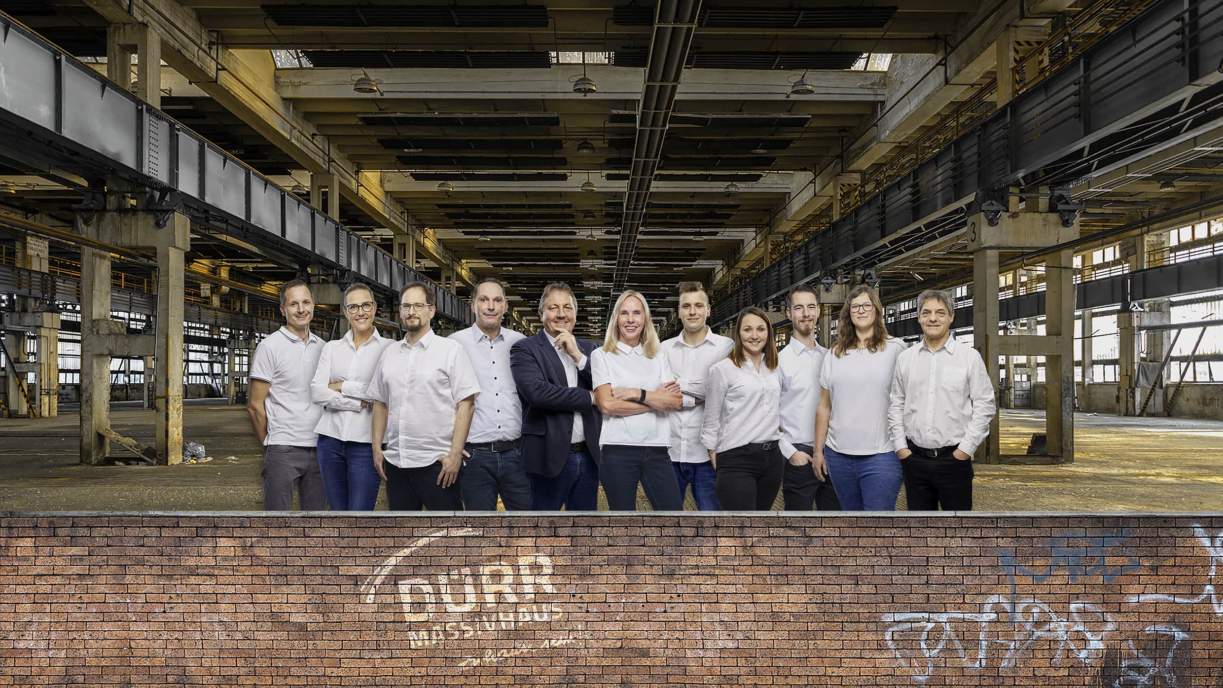 Gruppenfoto von 14 lächelnden Personen in weißer Kleidung vor einer industriellen Halle mit Backsteinmauer im Vordergrund und Dürr Musterdruck auf der Wand.
