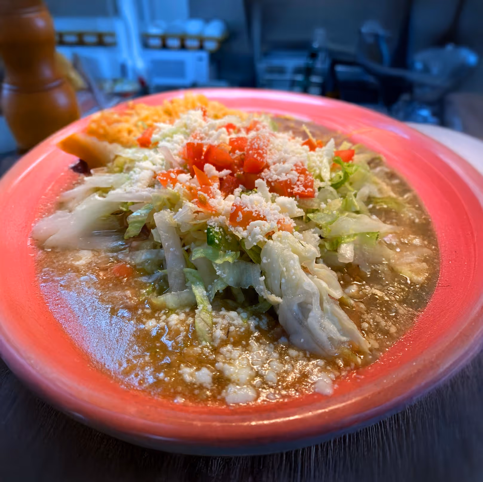 A vibrant pink bowl of steaming soup topped with shredded lettuce, diced tomatoes, and crumbled cheese, alongside a slice of bread, on a dark table.