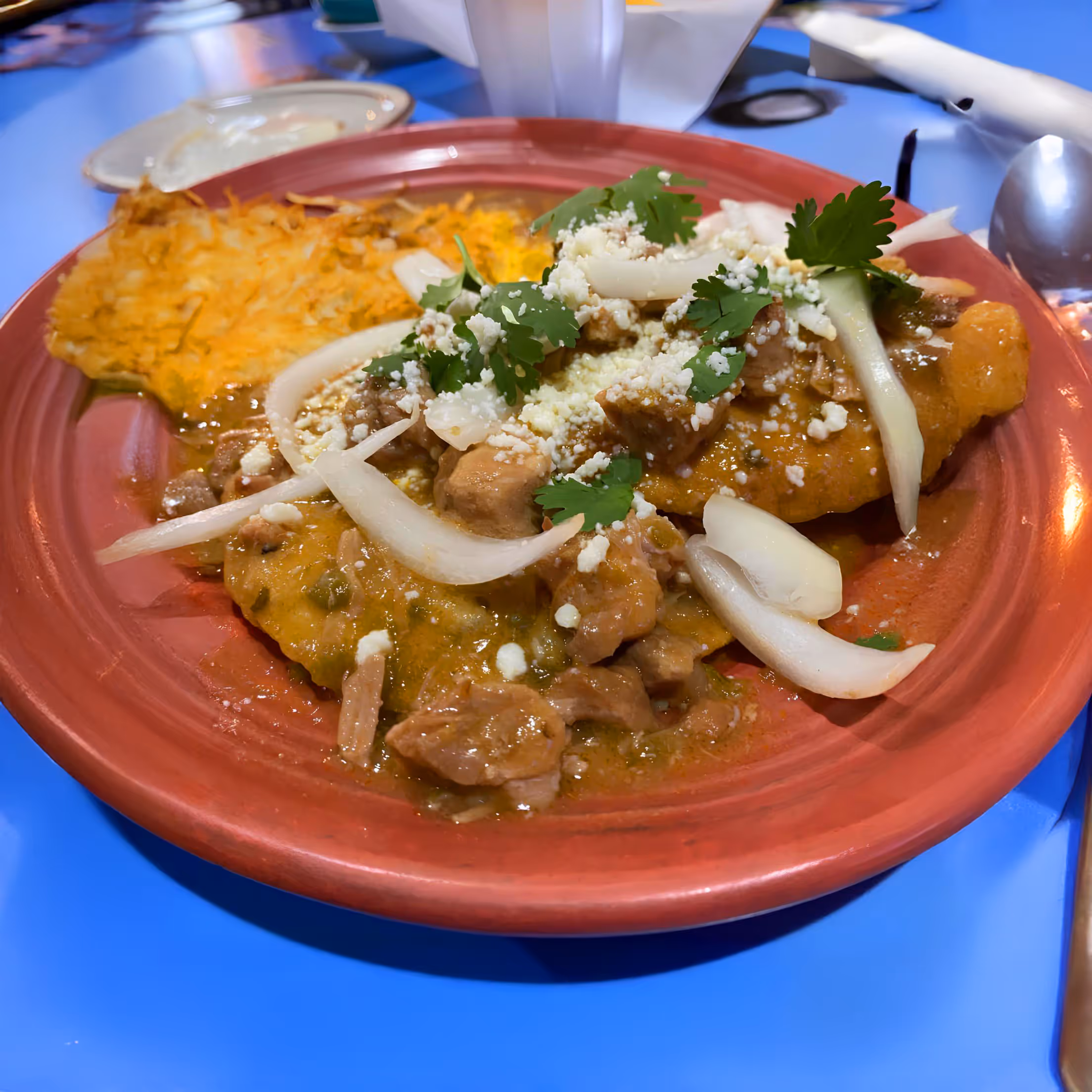 A vibrant red plate featuring a savory dish topped with cooked meat, onions, cilantro, and cheese. Beside it is a golden hash brown. 