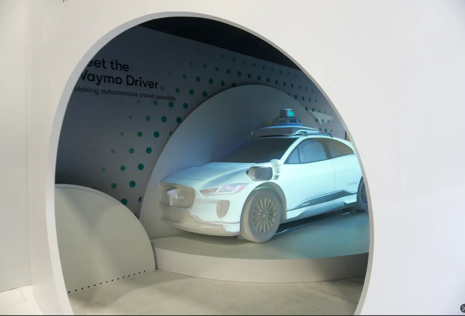 A futuristic Waymo exhibition display featuring a white Jaguar I-PACE autonomous vehicle positioned inside a sleek, circular white portal with dot-pattern graphics and 'Meet the Waymo Driver' branding.