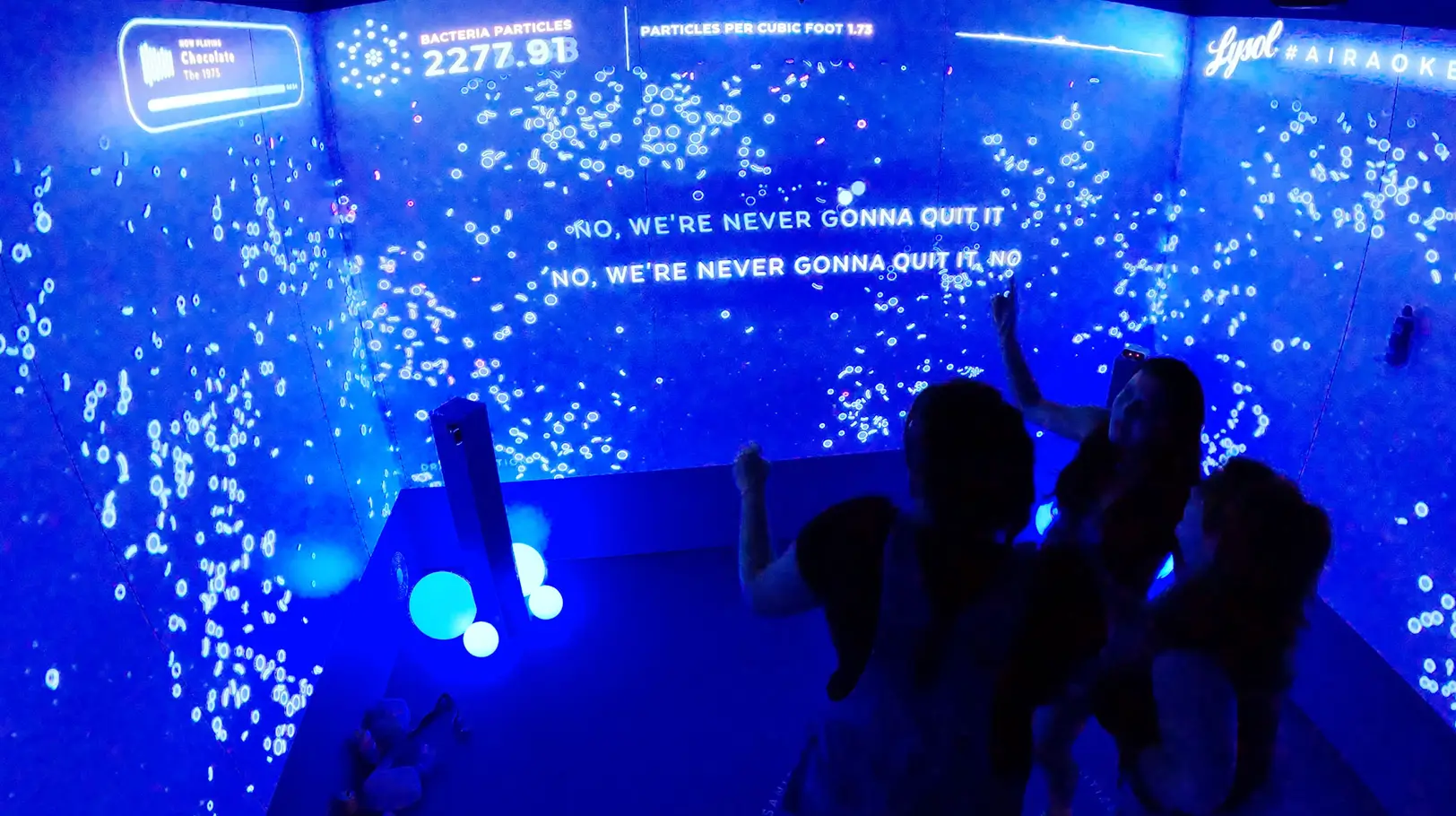 An immersive Lysol 'Air-aoke' activation featuring a dark room with floor-to-ceiling blue neon digital displays. The screens show floating white particles, bacteria counts, and song lyrics while participants interact with the multi-sensory environment.