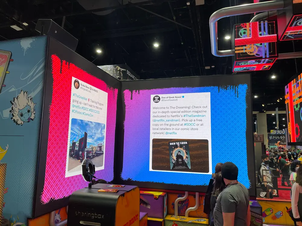 Two large digital screens displaying tweets about Netflix’s The Sandman at a convention, with people observing them.
