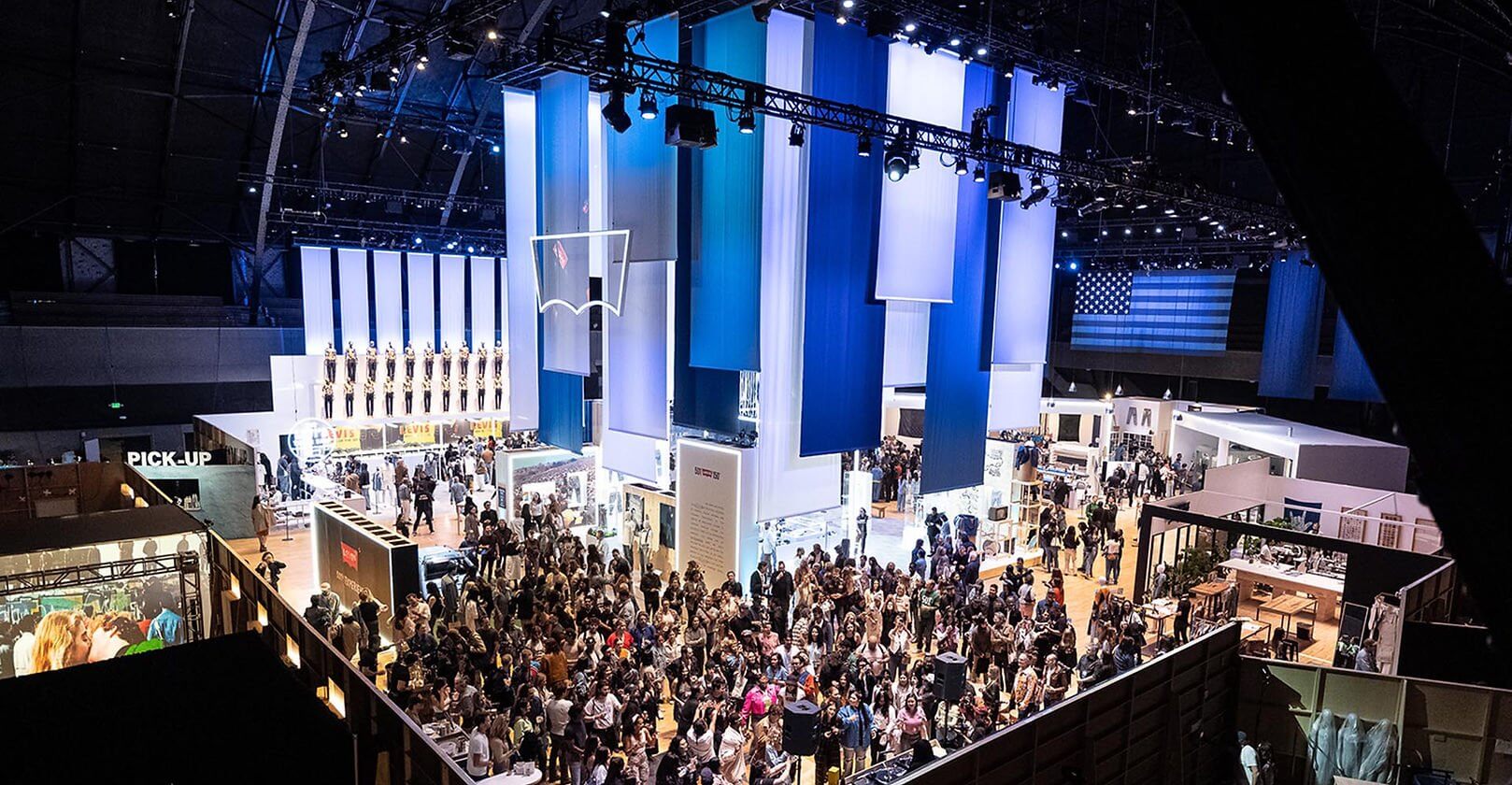 A wide, high-angle view of a massive Levi's brand activation in a large stadium, featuring towering vertical banners in shades of blue and white, a suspended glowing Levi's batwing logo, and a bustling crowd exploring various interactive retail and exhibit zones."