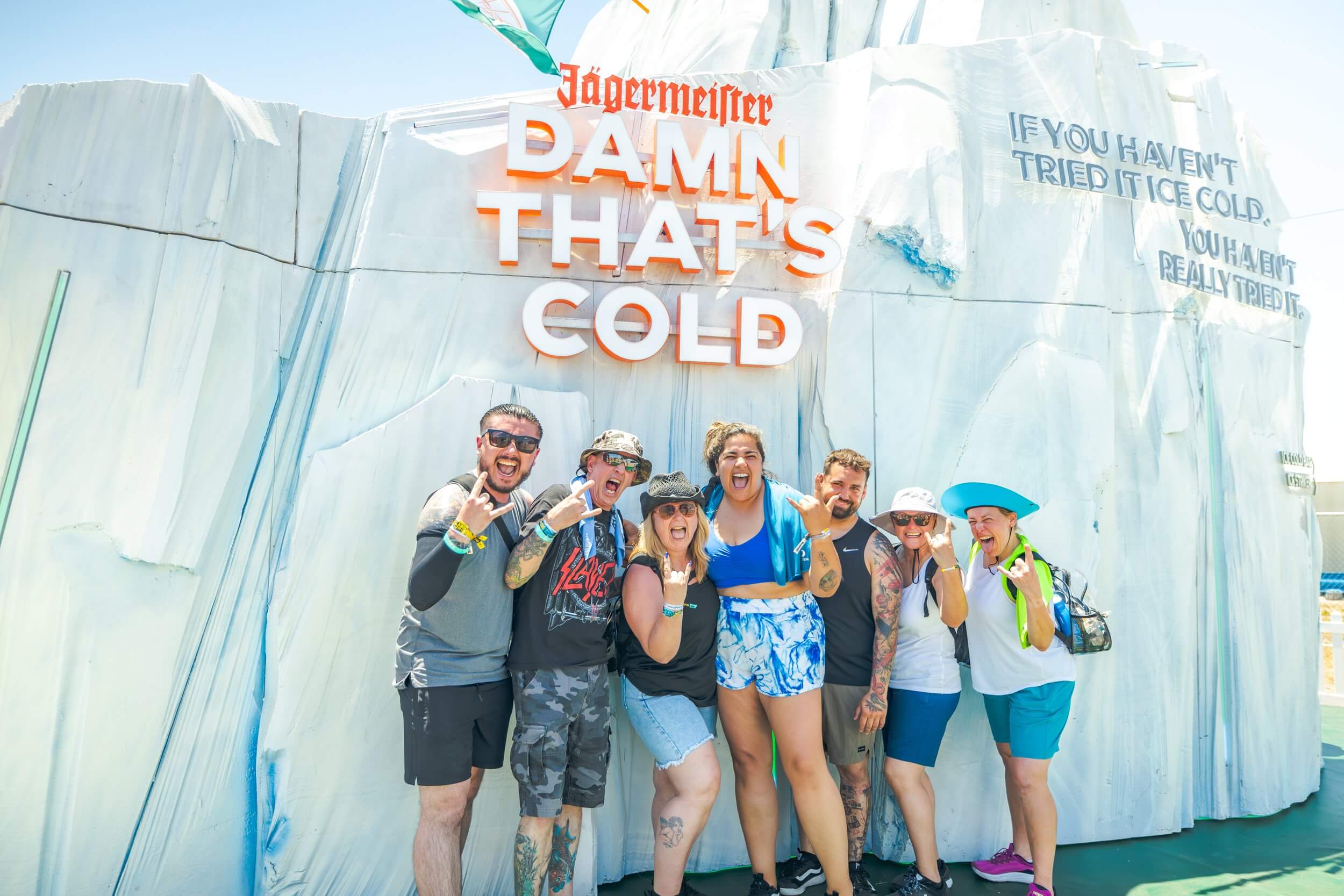 A group of cheerful festival-goers posing in front of a massive Jägermeister 'iceberg' installation. The textured white structure features the bold orange slogan 'DAMN THAT’S COLD' and branded Jägermeister signage under bright outdoor lighting.
