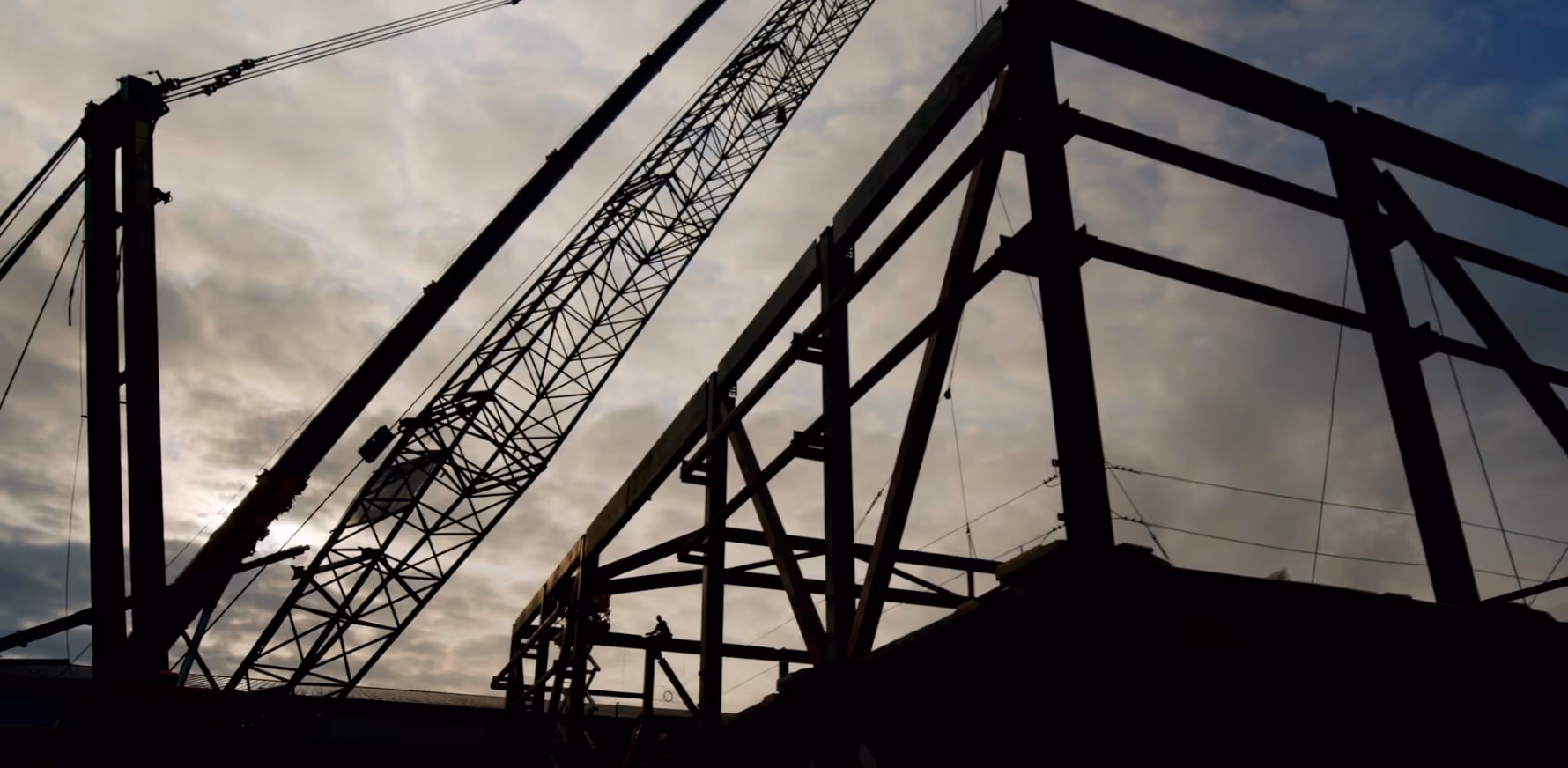 Silhouetted steel framework and crane at a construction site against a cloudy sky.