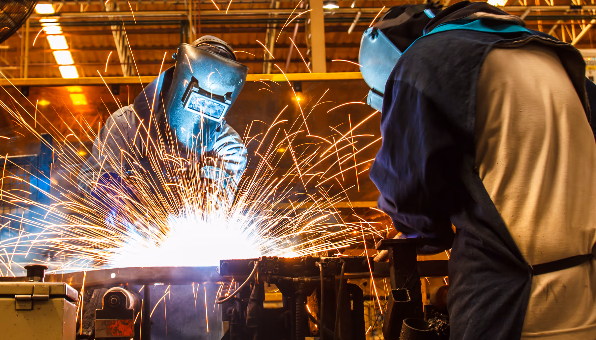 Two welders wearing protective helmets working with bright sparks flying in an industrial workshop.