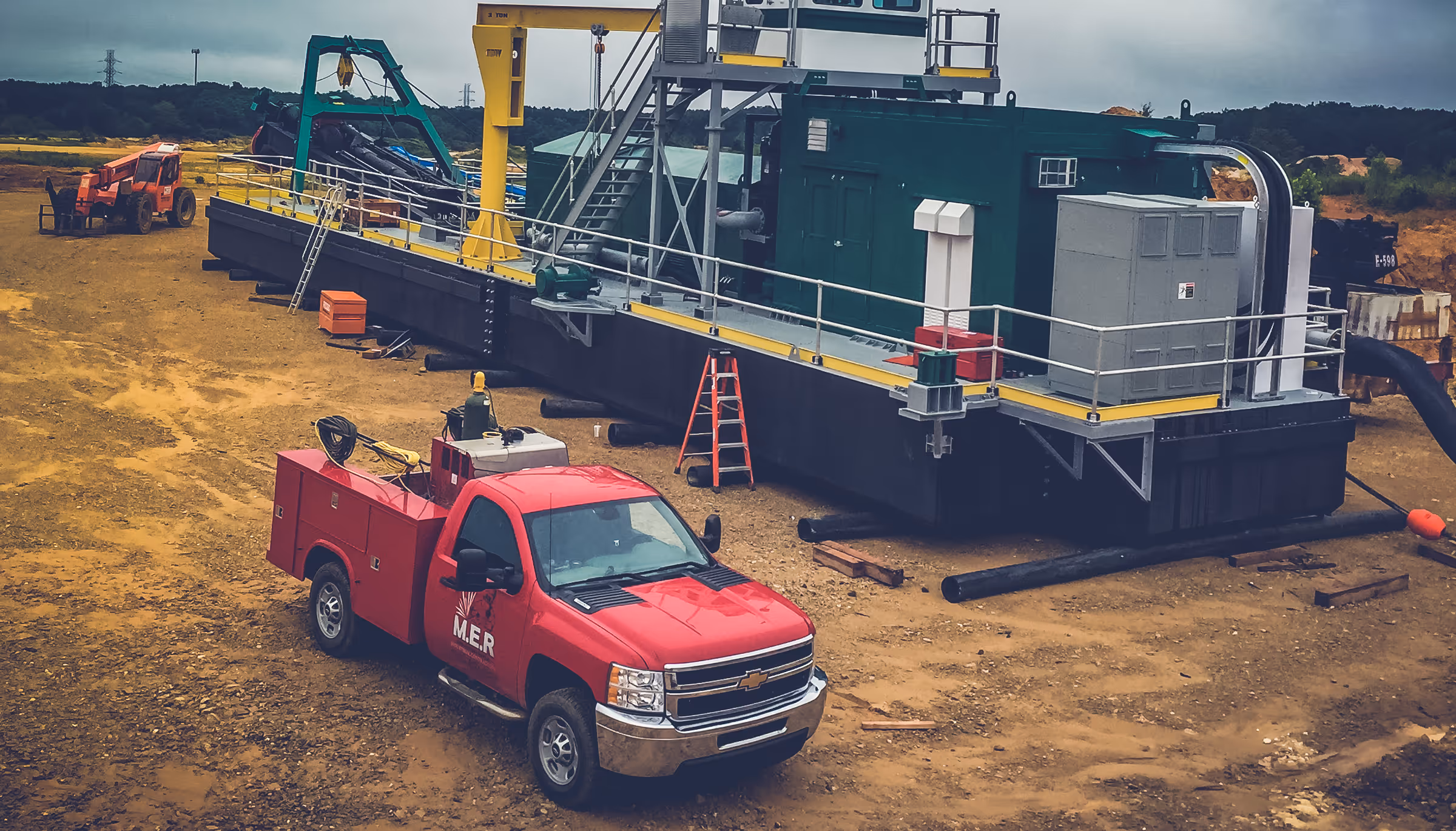 Red utility truck with M.E.R logo parked on a dirt site next to large industrial equipment and a yellow crane.
