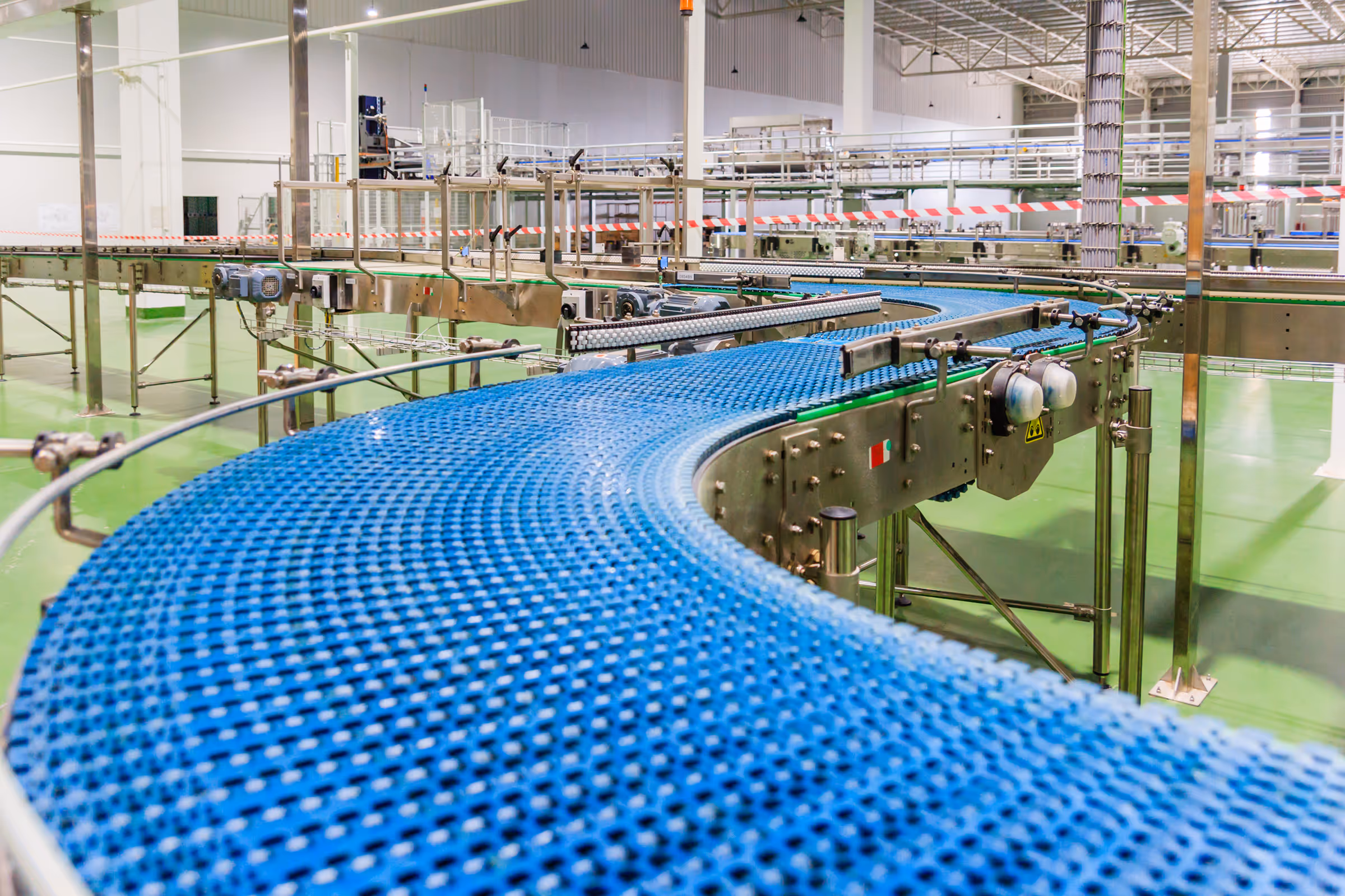 Curved blue industrial conveyor belt in an empty factory with green floors and metal structures.