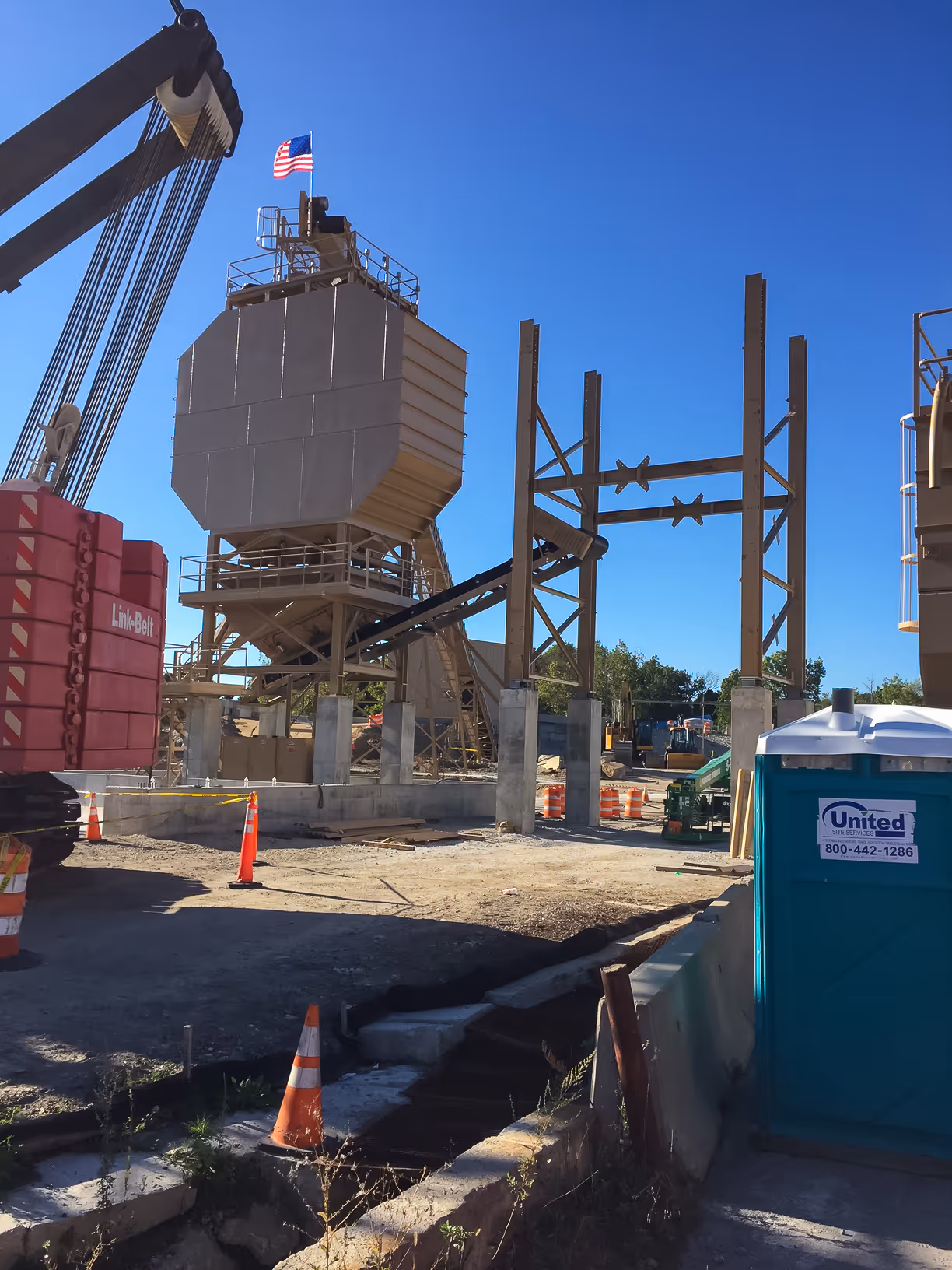 Construction site with industrial equipment, steel framework, an American flag, orange cones, and a blue portable toilet.