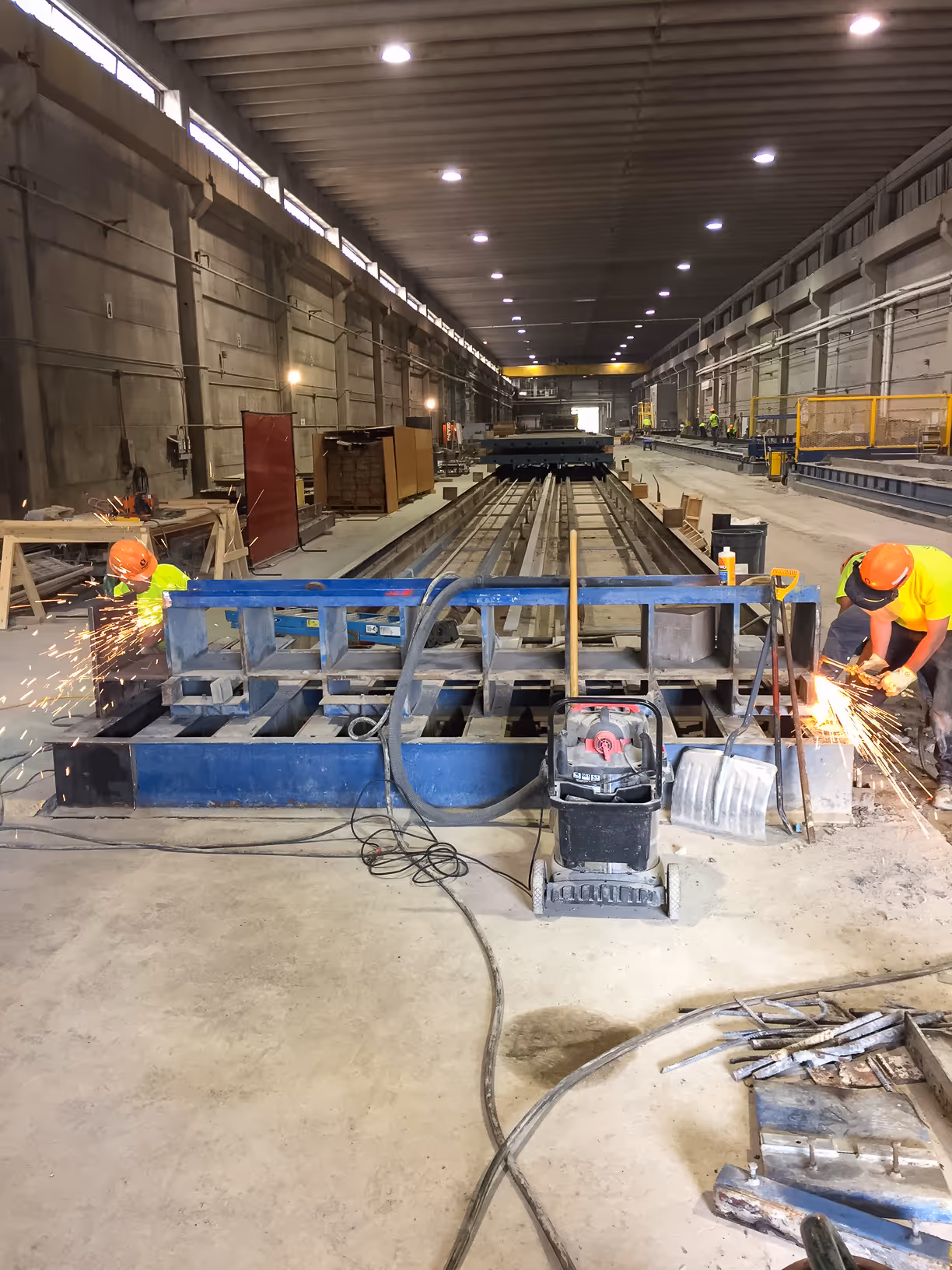Two construction workers wearing orange helmets grinding metal with sparks flying inside a large industrial workshop.