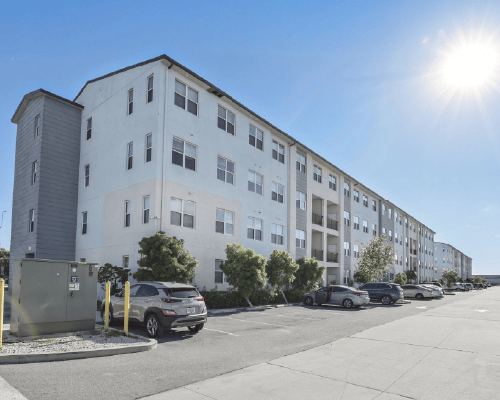 Long white apartment building with multiple floors, parked cars in front, under a bright sunny sky.