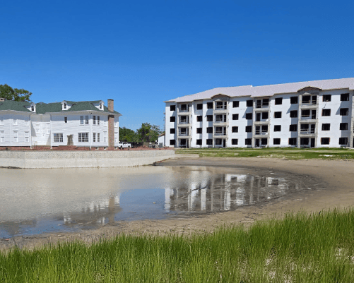 Two large white buildings near a shallow, sandy pond with grass in the foreground under a clear blue sky.
