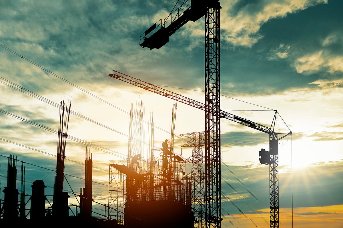 Silhouetted construction workers and cranes at a building site during sunset with a cloudy sky.
