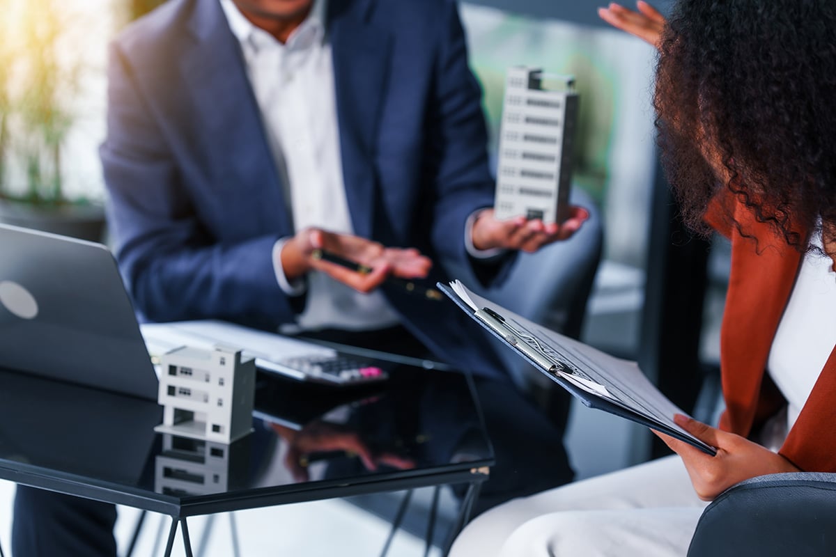 Two people discussing real estate with miniature building models on a glass table, one holding a clipboard and the other a scale model building.