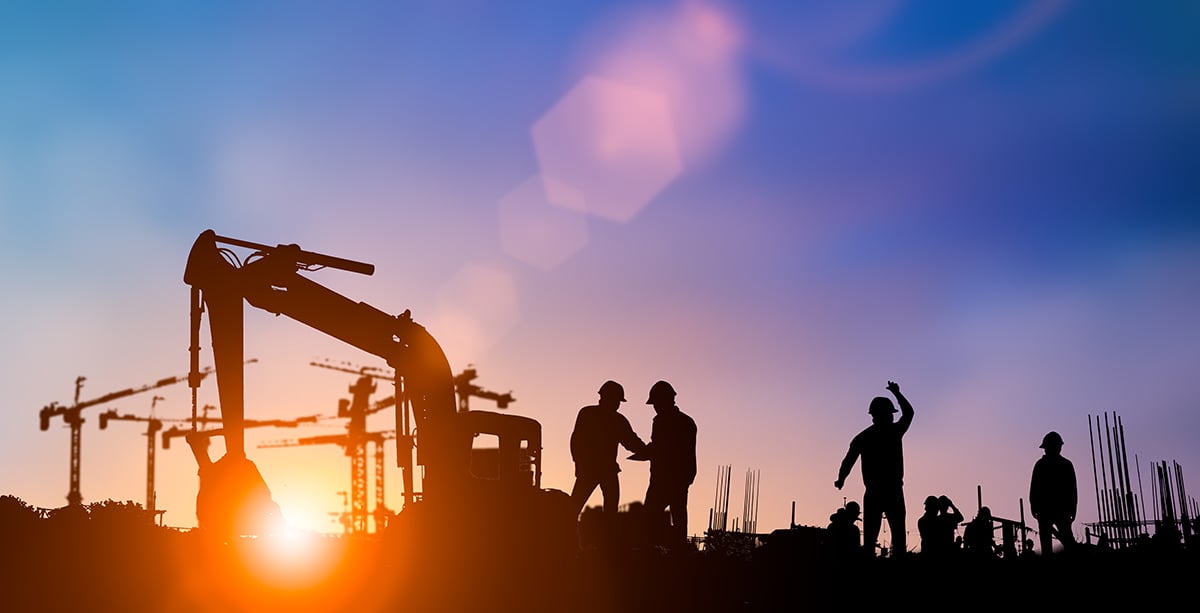 Silhouettes of construction workers and machinery with cranes at a construction site during sunset.