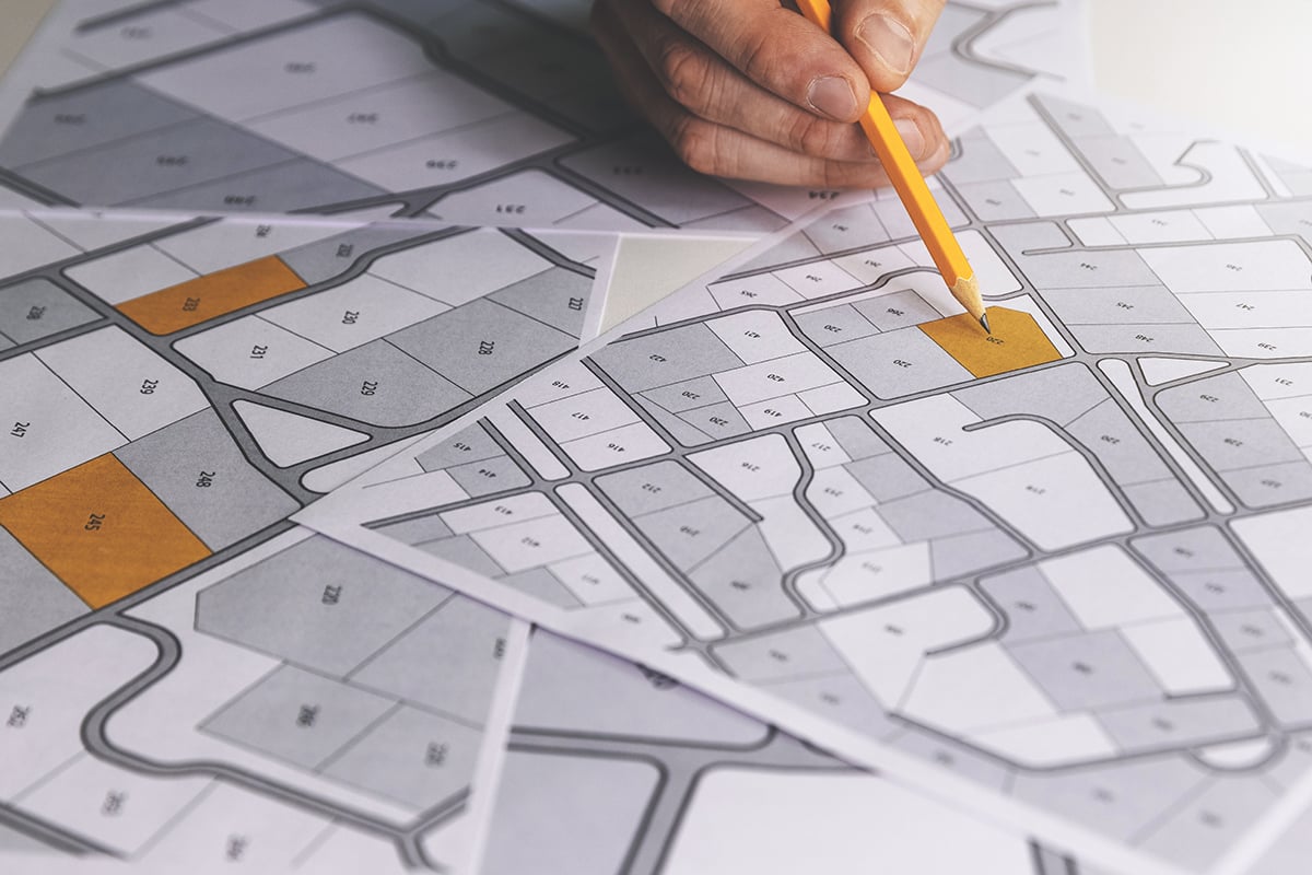 Hand holding a yellow pencil pointing to a highlighted plot on a residential land map with numbered parcels.