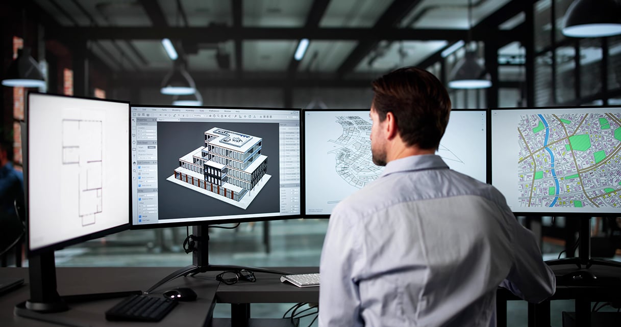 Person working at a desk with three monitors displaying architectural 3D model, floor plan, and city map.