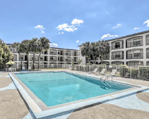 Outdoor swimming pool with clear blue water surrounded by a three-story white apartment building and palm trees under a partly cloudy sky.