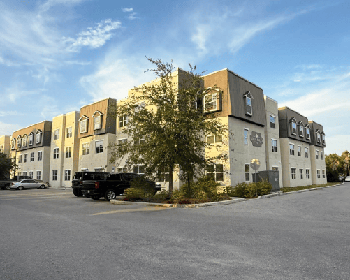 Three-story beige and brown apartment building with parked cars and a tree in front under a partly cloudy sky.