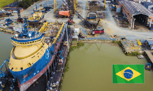 A large blue and yellow ship docked in a shipyard with cranes and industrial equipment, next to a body of water; Brazilian flag overlay at lower right.