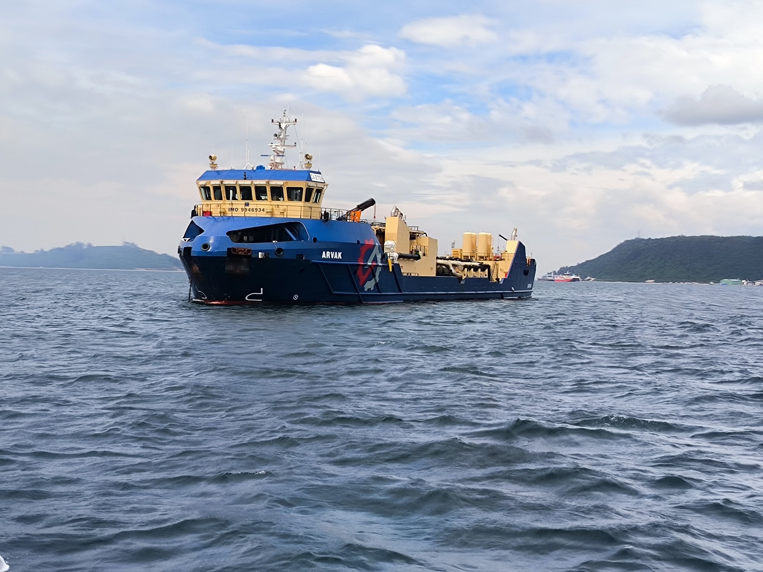 Blue and yellow industrial ship named ARVAK floating on calm sea with green hills and cloudy sky in the background.