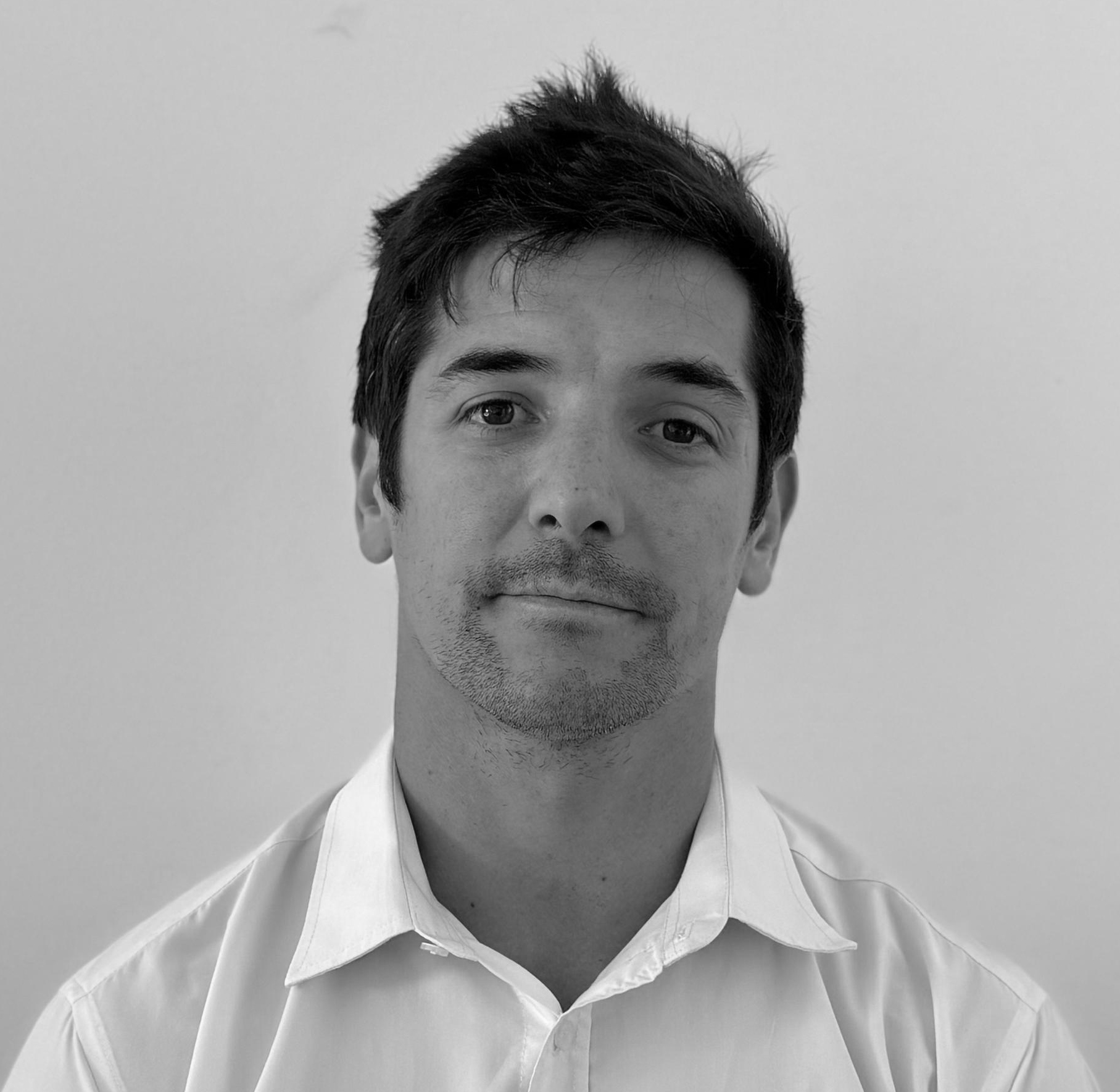 Portrait of a man with short dark hair and light stubble wearing a white collared shirt against a plain background.