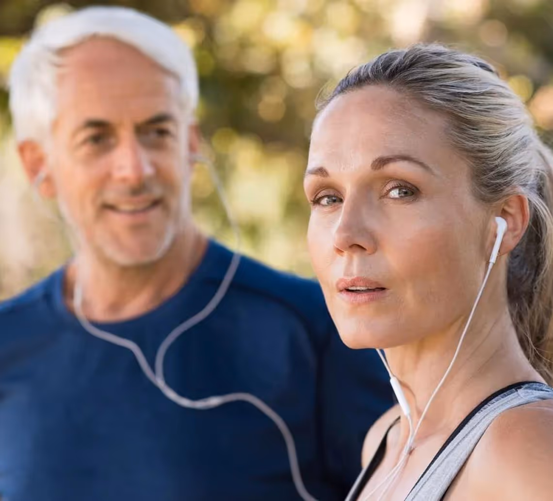 Mature woman and man outdoors wearing earphones, appearing to exercise or walk.