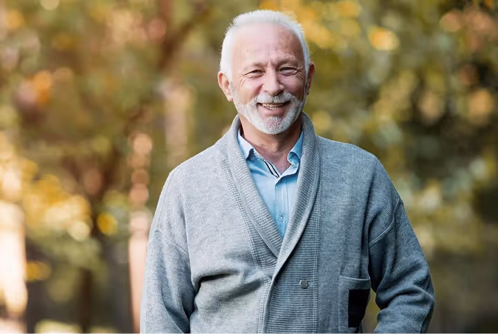 Smiling elderly man with white hair and beard wearing a gray cardigan and blue shirt outdoors.