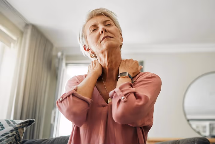 Older woman with short gray hair wearing a pink blouse, rubbing her neck with closed eyes in a living room.