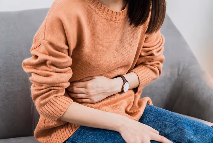Person sitting on a gray sofa holding their stomach, wearing a peach sweater and blue jeans.