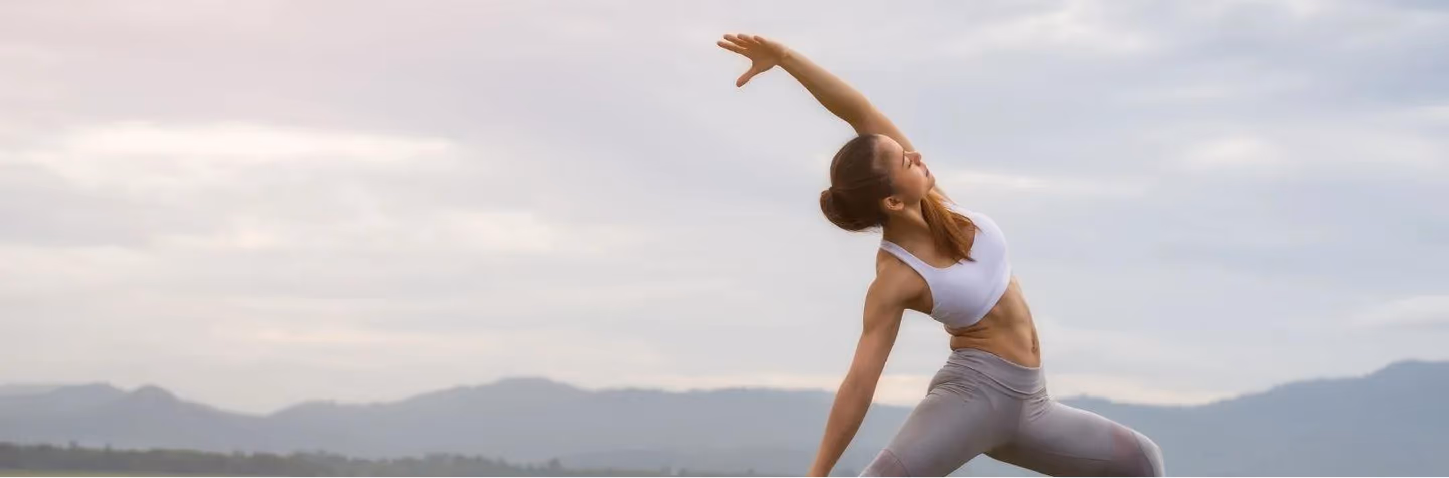 Woman in white sports bra and gray leggings stretching with one arm raised against a cloudy sky and distant mountains.