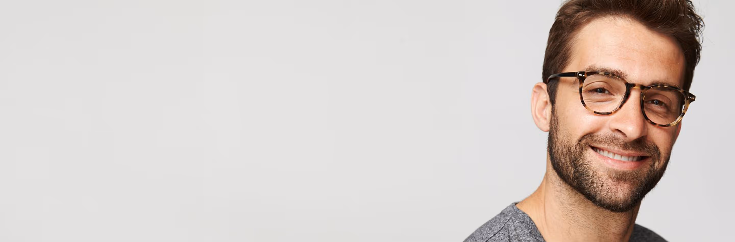 Smiling man with brown hair, beard, and tortoiseshell glasses wearing a grey shirt against a plain light background.