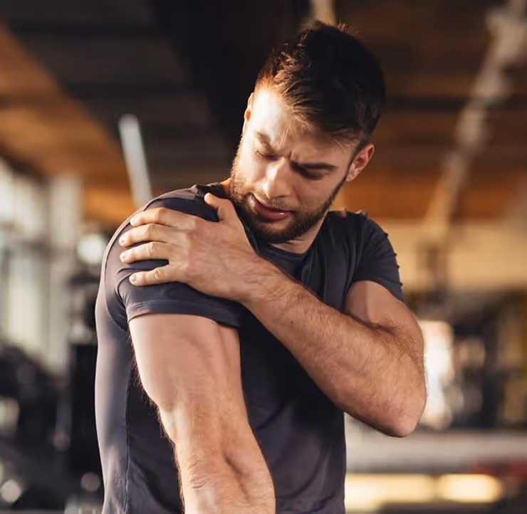 Man in gym wearing a dark t-shirt holding his shoulder in pain.