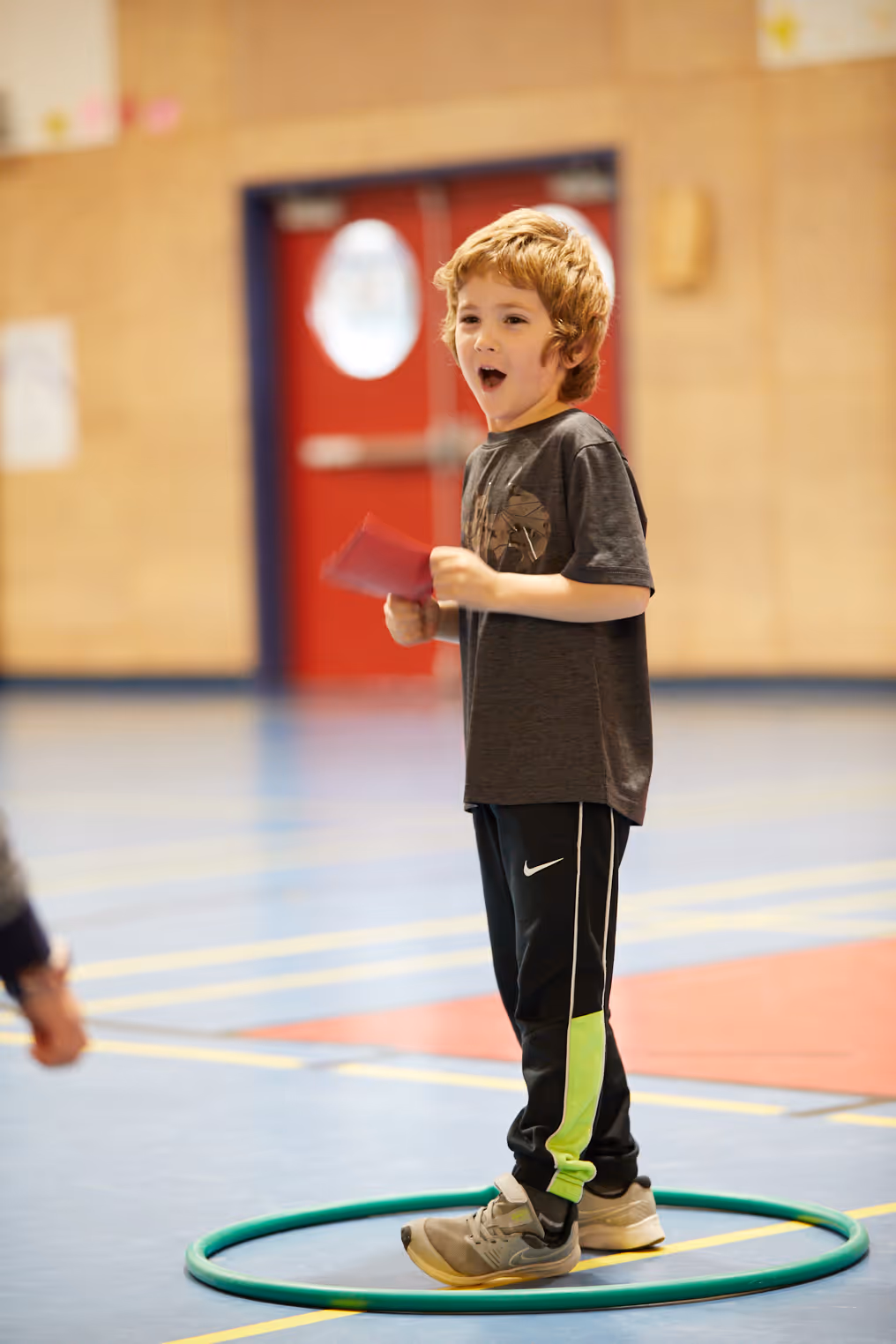Young boy standing inside a green hoop on a gym floor, holding a red card and speaking with an open mouth.