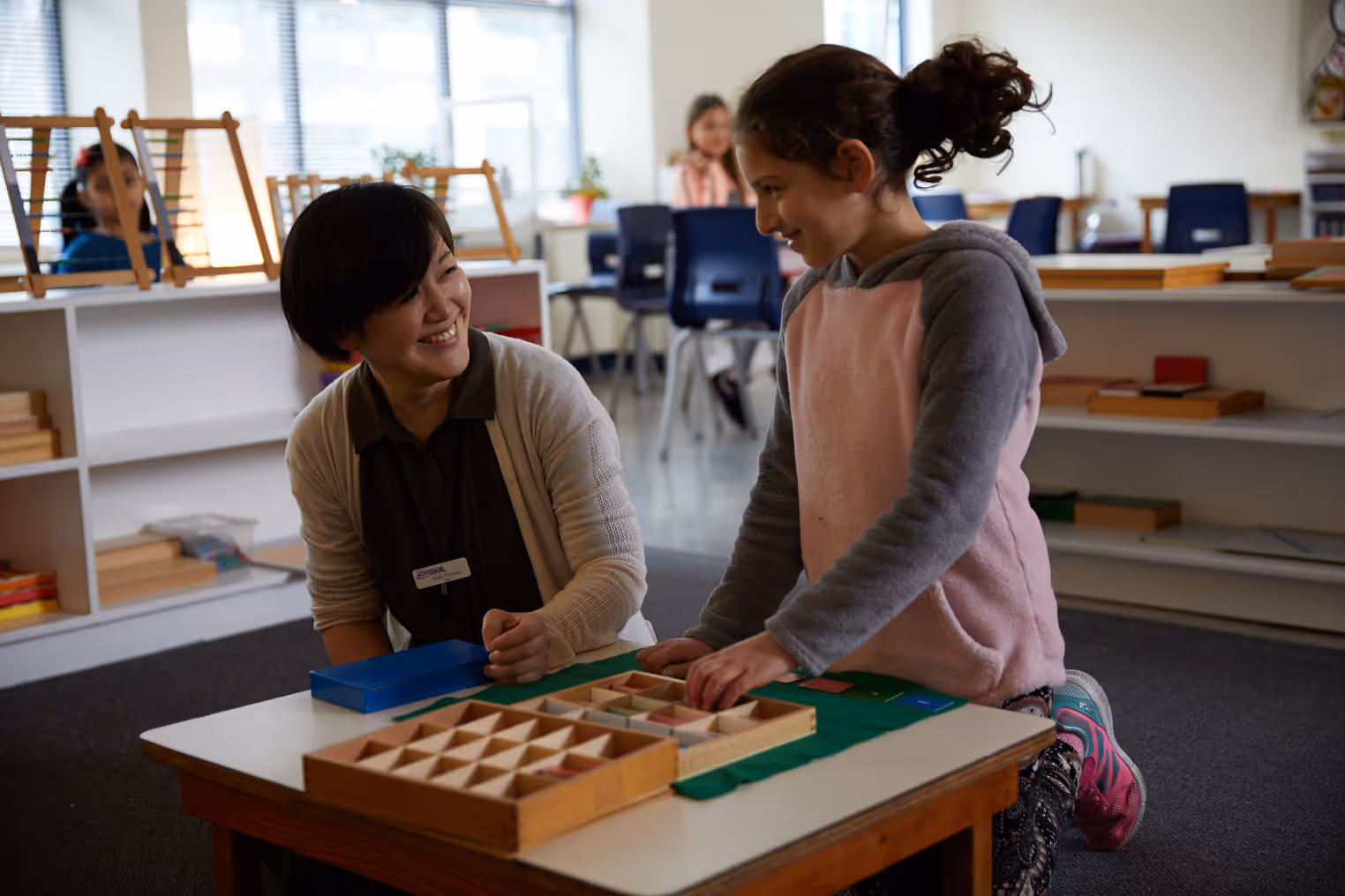 Teacher and young student smiling and working with educational materials at a table in a classroom.