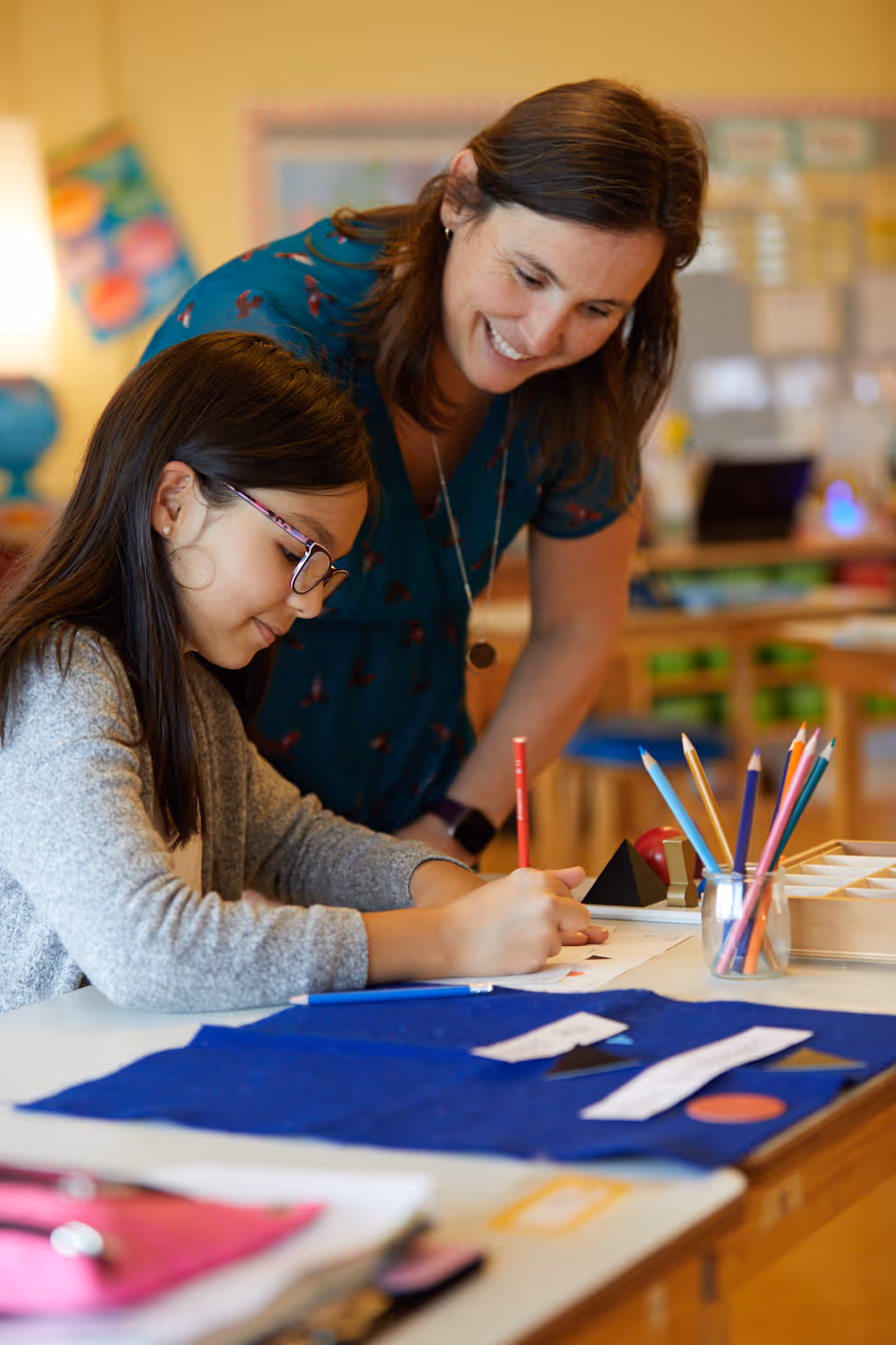 Teacher smiling and leaning over a student who is writing with a pencil in a classroom.