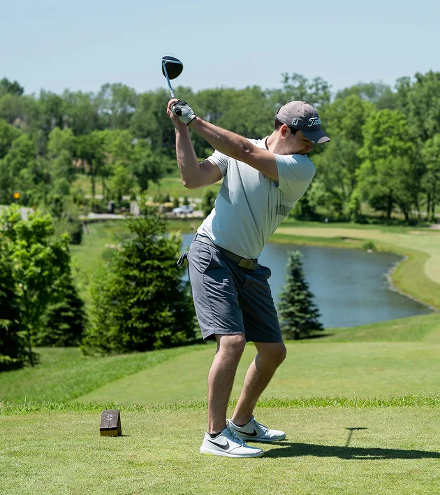 Man in a gray cap and shorts mid-swing on a golf course near a water hazard and trees.