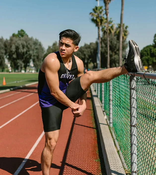 Young male athlete in black and purple uniform stretching his leg on a chain-link fence beside a running track under clear sky.