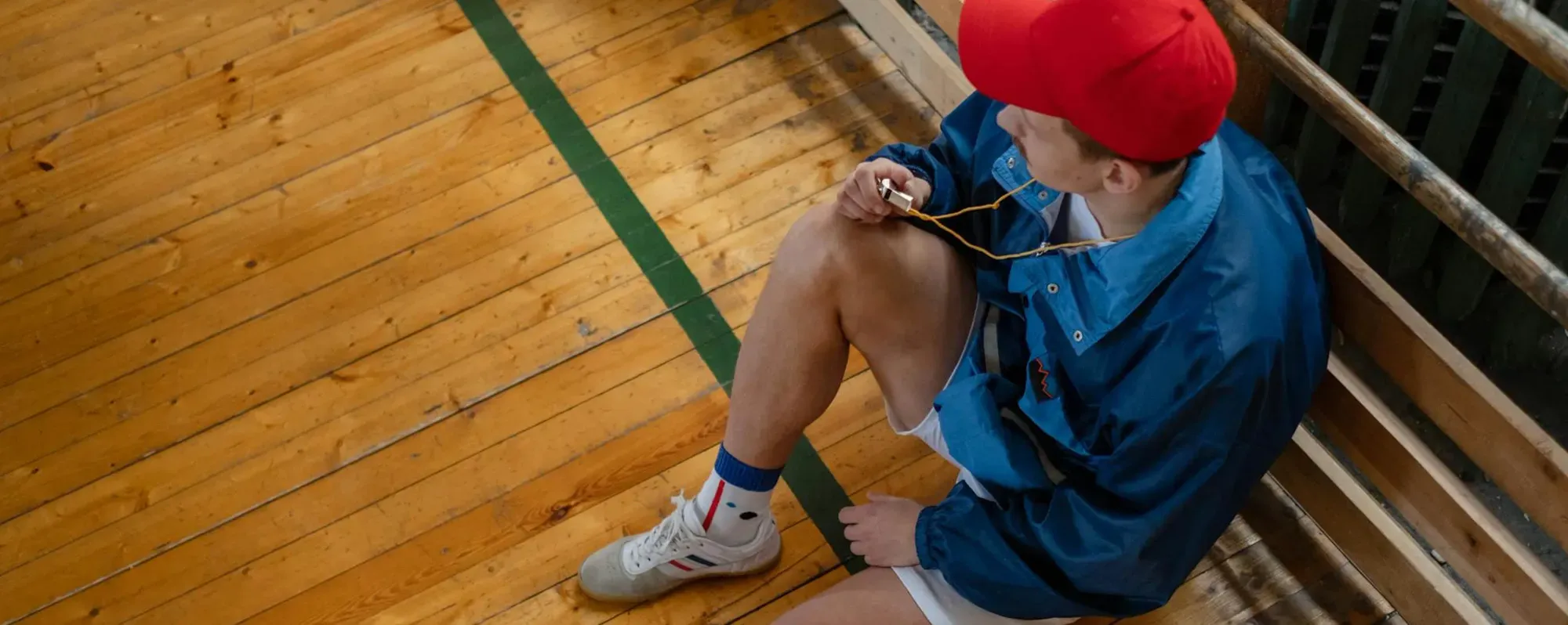 Young man in a red cap and blue jacket sitting on a wooden bench in a gymnasium holding a whistle on a yellow lanyard.